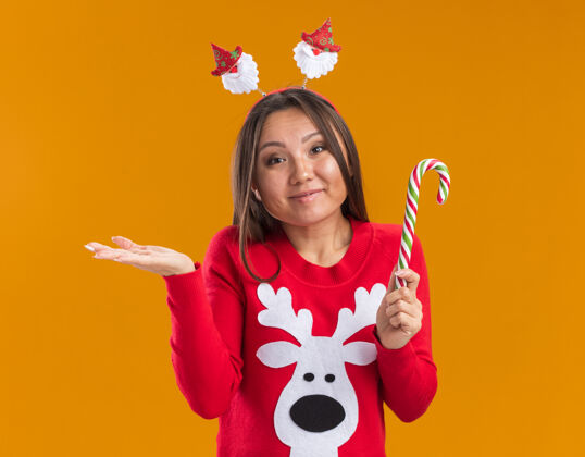 年轻人高兴的年轻亚洲女孩戴着圣诞发箍和毛衣拿着圣诞糖果摊开手孤立的橙色背景抱蔓延糖果