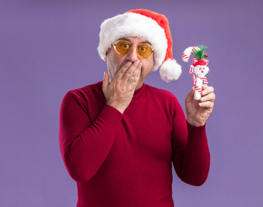 帽子中年男子戴着圣诞帽戴着黄色眼镜拿着圣诞糖果手杖看着摄像机惊讶地捂着嘴站在紫色的背景上糖果圣诞老人封面