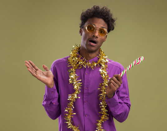 年轻令人印象深刻的年轻非裔美国人戴着眼镜 脖子上戴着金属丝花环 手持圣诞糖果手杖 看着相机 显示出橄榄绿背景上孤立的空手橄榄糖果空
