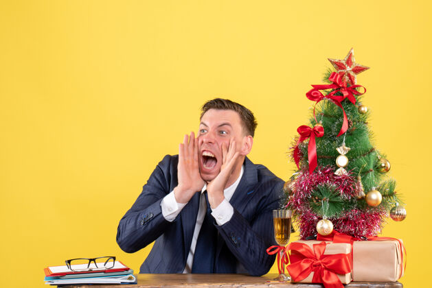 沟通者愤怒的男人坐在圣诞树旁的桌子前大声喊叫 黄色的墙上摆着礼物树男人愤怒