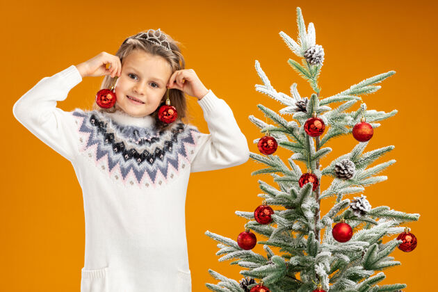 脖子微笑的小女孩站在圣诞树旁 戴着头饰 脖子上戴着花环 耳朵上挂着圣诞球 背景是橙色的树微笑球