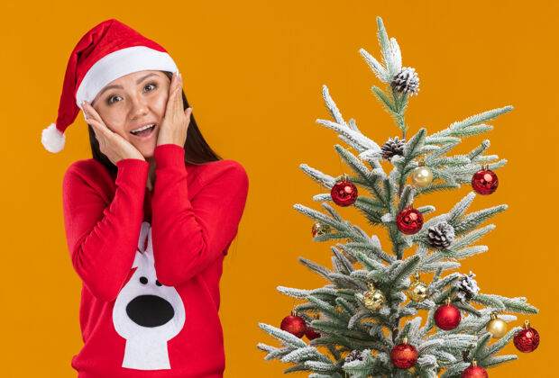 脸颊微笑的亚洲女孩戴着圣诞帽 穿着毛衣站在圣诞树旁 双手放在橘色背景下孤立的脸颊上帽子女孩圣诞节