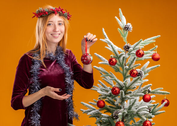 舞会微笑着站在圣诞树旁的年轻漂亮女孩身穿红色连衣裙 脖子上戴着花环 手里拿着橙色背景上孤立的圣诞球女孩圣诞节衣服