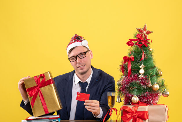 商人商务人士手持卡片和圣诞礼物坐在圣诞树旁的桌子前 黄色的礼物礼物黄色坐着