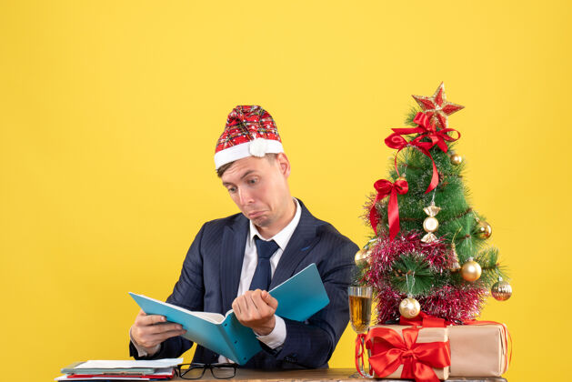 商务人士商务人员坐在圣诞树旁的桌子旁检查文件和黄色墙上的礼物的前视图公司人商人