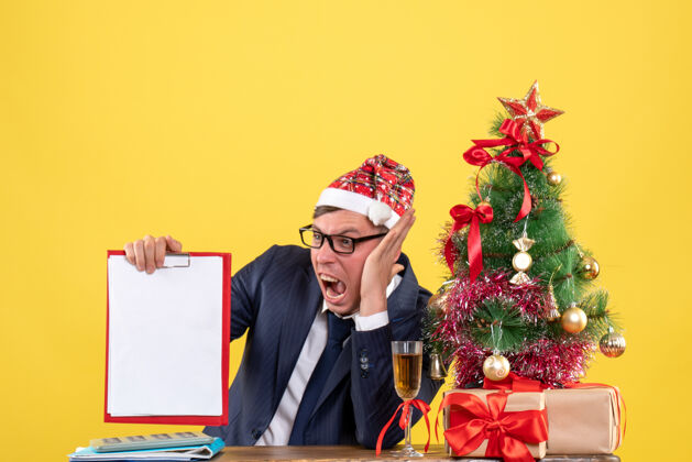 愤怒愤怒的商人坐在圣诞树旁的桌子前 黄色的墙上摆着礼物愤怒的商人经理办公室