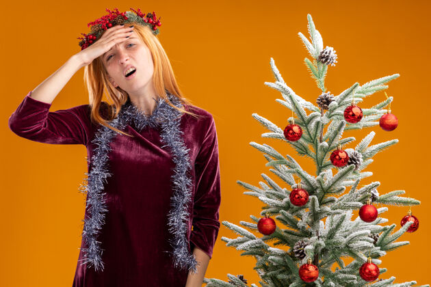 花环疲惫的年轻漂亮女孩站在圣诞树旁 身穿红色连衣裙 脖子上戴着花环 手前额孤立在橙色背景上圣诞节前额衣服