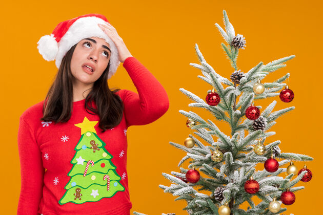 烦恼戴着圣诞帽的年轻白人女孩生气地把手放在额头上 眼睛滚动着站在橙色背景下的圣诞树旁 还有复制空间女孩年轻放