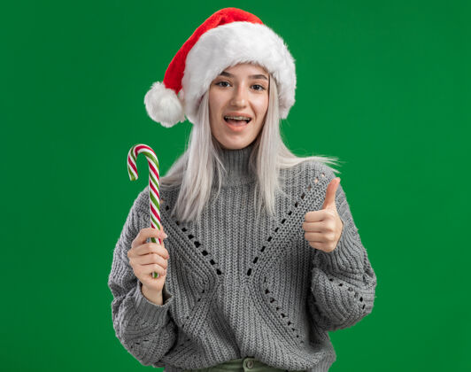 年轻人快乐的金发女郎 穿着冬衣 戴着圣诞帽 手里拿着糖果手杖 站在绿色的背景下 看着镜头 开心地微笑着手杖糖果圣诞老人