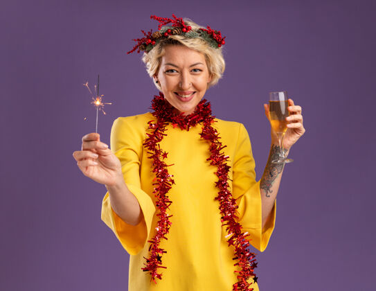 花环微笑的金发女郎戴着圣诞花环 脖子上戴着金箔花环 手里拿着节日火花和香槟 看着紫色背景上孤立的摄像机杯子香槟金发