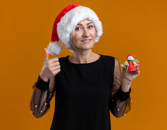 中年微笑的中年金发女人戴着圣诞帽看着摄像机向镜头展示小雪人雕像 并在橙色背景上孤立地竖起大拇指小圣诞节显示