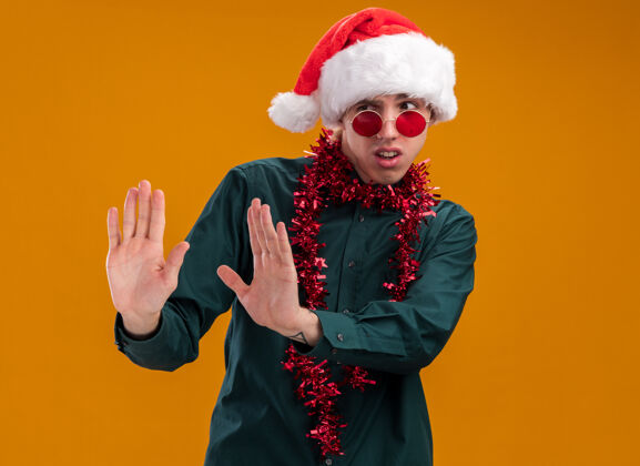 圣诞老人戴着圣诞帽戴着眼镜 脖子上戴着金箔花环的年轻金发男子 在橙色背景上做着拒绝的手势 并留有复印空间年轻眼镜花环