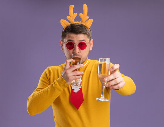 站着穿着黄色高领毛衣 戴着红色眼镜的年轻人 打着有趣的红色领带 戴着鹿角边 手里拿着两杯香槟 快乐而积极地站在紫色的背景上年轻角眼镜