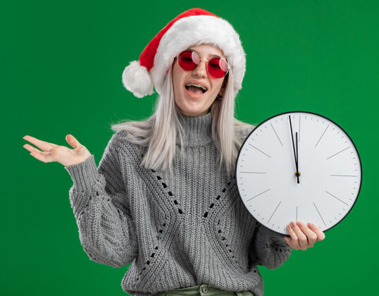 毛衣年轻的金发女郎穿着冬衣 戴着圣诞帽 戴着红眼镜 拿着挂钟 站在绿色的背景下 快乐而兴奋地看着摄像机金发抱着年轻人