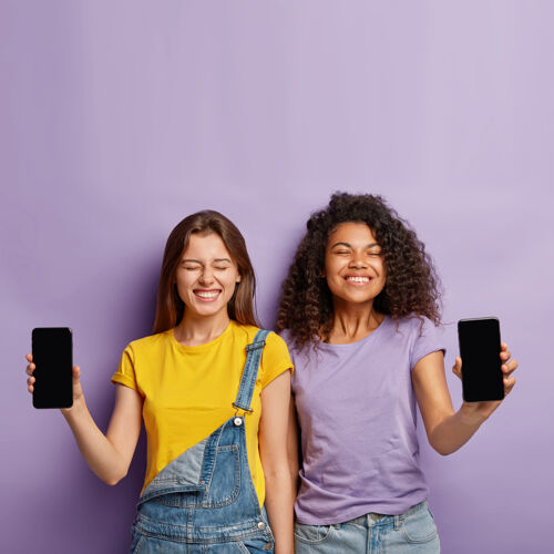 应用程序快乐多样的姐妹们站在一起 展示空白屏幕的手机 拥有积极的外表 为新产品做广告空互联网手持