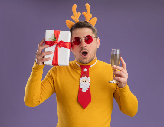 惊讶一个穿着黄色高领毛衣 戴着红色眼镜的年轻人 打着有趣的红色领带 戴着鹿角边 手里拿着一杯香槟 站在紫色的背景下 他看上去既惊讶又惊奇拿着边缘杯子