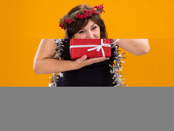 中年高兴的中年妇女头戴圣诞花环 脖子上戴着金箔花环 手里拿着礼包 看着隔离在橙色背景上的相机包花环手捧
