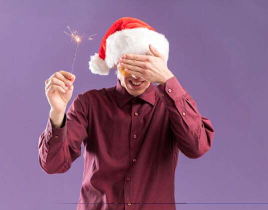 圣诞老人微笑着的年轻金发男子戴着圣诞帽 戴着圣诞眼镜 手里拿着节日用的闪闪发光的东西 用手遮住眼睛 在紫色的背景上隔离开来眼镜眼睛金发