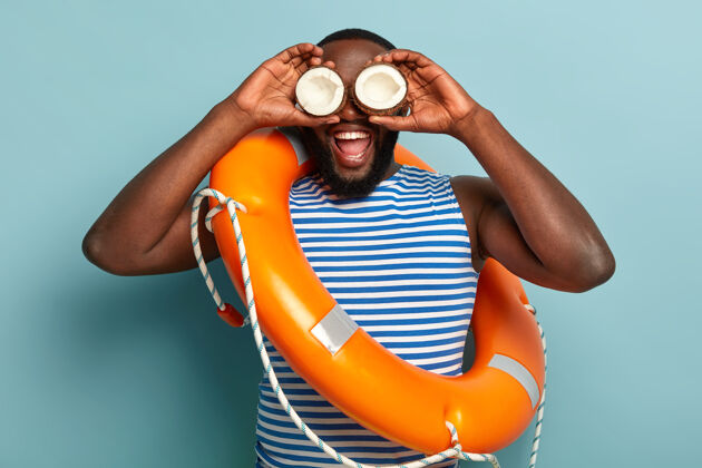 戒指有趣的黑皮肤男人独自一人玩 拿着椰子在眼睛上 试着看向海滩的远处人类游泳助理