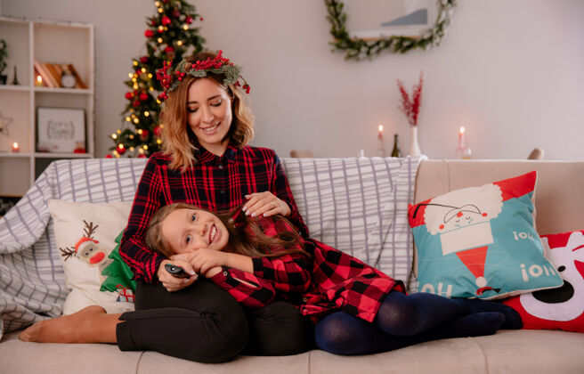 大腿高兴的妈妈拿着冬青花环 拿着电视遥控器 看着躺在大腿上的女儿坐在沙发上 在家里享受圣诞节时光女儿家沙发