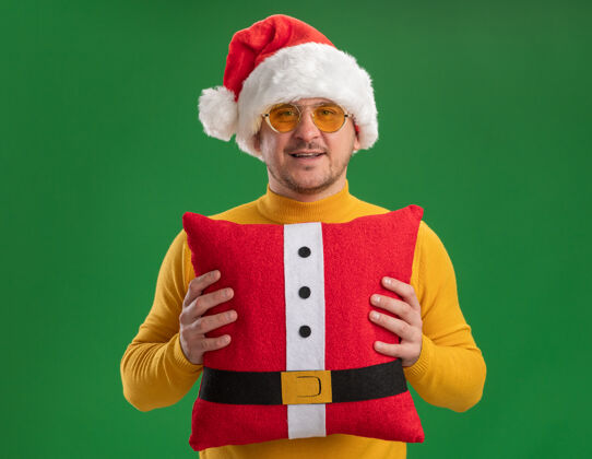 高领毛衣快乐的年轻人穿着黄色高领毛衣 戴着圣诞帽 戴着眼镜 抱着红色滑稽的枕头 站在绿色的背景下 面带微笑地看着相机眼镜圣诞老人帽子
