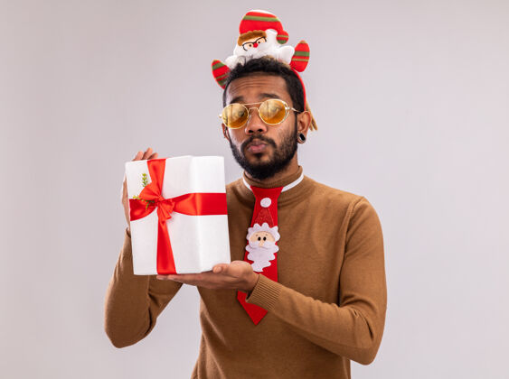 抱着一个穿着棕色毛衣 头戴圣诞老人戒指 打着有趣的红色领带的非裔美国人拿着一件礼物 站在白色背景下 看上去很困惑困惑轮辋美国人