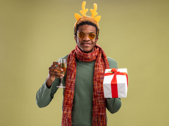 美国快乐的非洲裔美国人 戴着有趣的边 戴着鹿角 脖子上围着围巾 手里拿着一杯香槟和圣诞礼物 站在绿色的背景下 看着相机 微笑着 自信地站着鹿微笑非洲