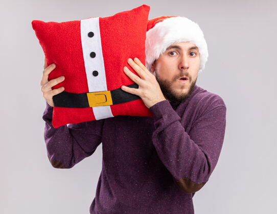 困惑一个穿着紫色毛衣 戴着圣诞帽的年轻人拿着一个滑稽的枕头 站在白色的背景下 困惑而担心地看着镜头搞笑站着担心