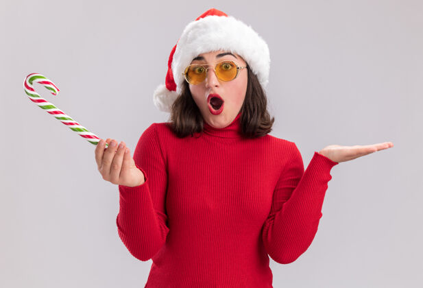 圣诞老人穿着红色毛衣 戴着圣诞帽 戴着眼镜 手持糖果手杖的年轻女孩看着镜头 惊讶而困惑地举起双臂站在白色背景上糖果举起来女孩