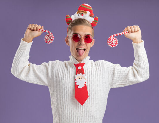 圣诞老人顽皮的年轻帅哥戴着圣诞老人的头带和领带戴着眼镜拿着圣诞糖果手杖看着相机显示舌头孤立在紫色背景上英俊眼镜手杖