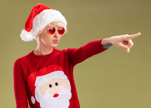 橄榄自信的金发女郎 戴着圣诞帽 穿着圣诞老人的圣诞毛衣 戴着眼镜 一边看着一边 噘着嘴唇 橄榄绿的背景上孤立着指向看自信