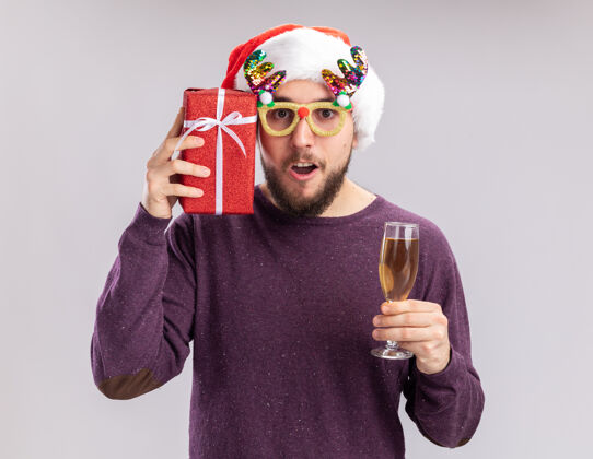 圣诞节身穿紫色毛衣 头戴圣诞帽 戴着滑稽眼镜 手持香槟的快乐年轻人 站在白色的墙上 微笑着向大家介绍新年假期的概念圣诞老人站着拿着