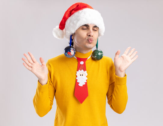 耳朵穿着黄色高领毛衣的快乐的年轻人和耳朵上戴着圣诞球的圣诞老人快乐有趣地闭着眼睛站在白色的背景上高领毛衣年轻搞笑