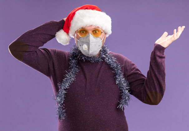 保持一个戴着圣诞老人帽 脖子上戴着金属丝花环 戴着眼镜和防护面具的中年男人看着镜头 手空着 手放在头后 在紫色背景下与世隔绝保护空脖子