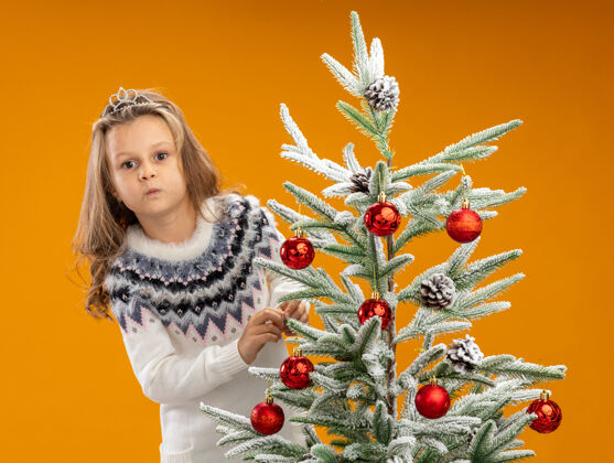 快乐高兴的小女孩站在圣诞树后面戴着头饰 脖子上的花环孤立在橙色的背景上脖子圣诞快乐站着