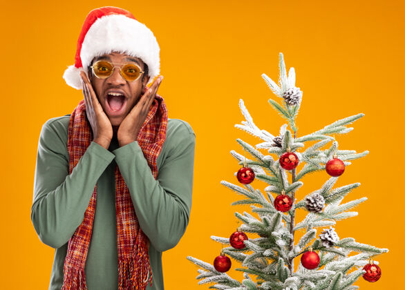 兴奋戴着圣诞帽 脖子上围着围巾的非洲裔美国人站在橙色背景下的圣诞树旁 高兴而兴奋地看着摄像机脖子圣诞老人圣诞节