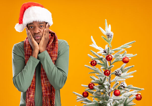脖子戴着圣诞帽 脖子上围着围巾的非裔美国人站在橙色背景下的圣诞树旁 看着摄像机 心烦意乱 忧心忡忡烦恼帽子圣诞老人