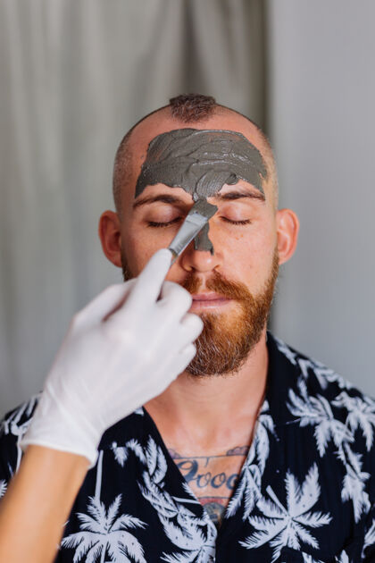 冷静美容美容师在美容诊所用刷子在年轻帅哥脸上涂泥面膜治疗粘土男人