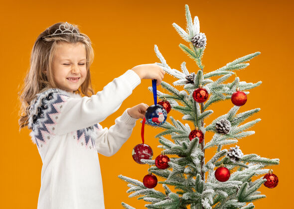 女孩高兴地闭着眼睛站在圣诞树旁的小女孩戴着头饰 脖子上戴着花环 手里拿着孤立在橙色背景上的圣诞球头饰站着高兴
