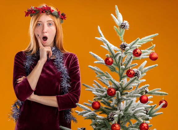 年轻害怕的年轻漂亮女孩站在圣诞树旁 穿着红色的连衣裙 脖子上戴着花环 手指放在橘黄色背景下的脸颊上衣服站着树