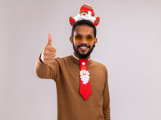 非洲一个身穿棕色毛衣 头戴圣诞老人耳环 打着有趣的红色领带的非裔美国人看着镜头 兴高采烈地微笑着 竖起大拇指站在白色背景上毛衣微笑领带
