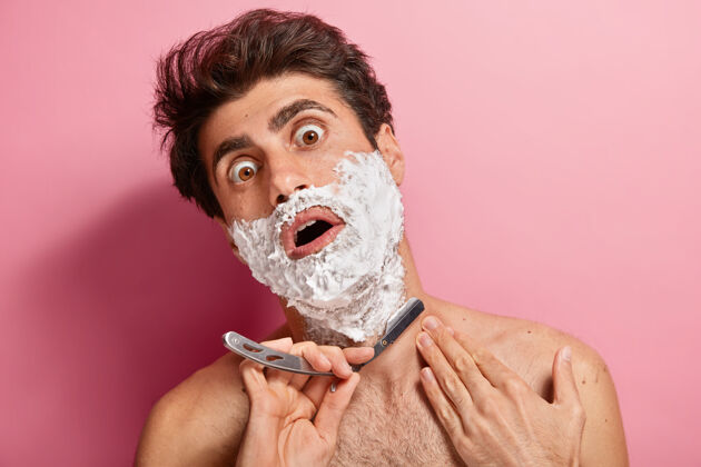 凝视震惊的年轻人涂上泡沫 准备修剪胡须 拿着剃须刀刀片 感觉每天刮胡子又厚又累皮肤剃须玫瑰色