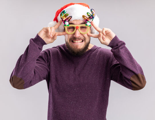 圣诞老人一个穿着紫色毛衣 戴着圣诞帽 戴着滑稽眼镜 站在白色背景上 眼睛附近有v字标志的快乐的年轻人眼镜搞笑年轻