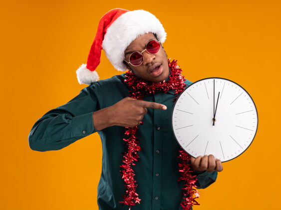 美国人戴着圣诞帽 戴着花环 戴着太阳镜 拿着时钟 食指指着时钟的非裔美国人站在橙色背景上非洲人拿着站着