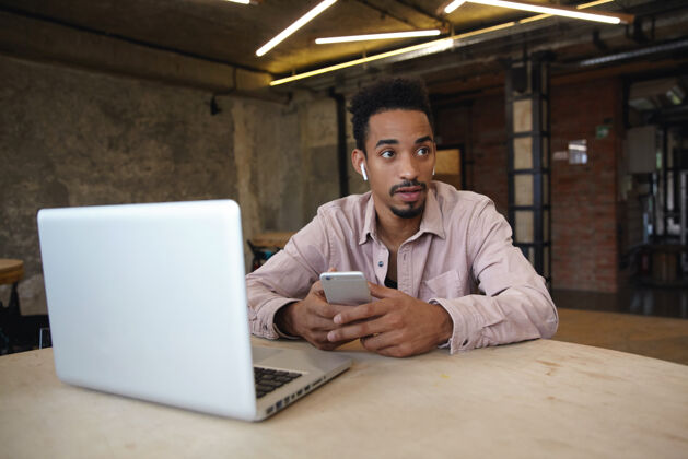 室内室内照片：年轻漂亮的黑皮肤男性 留着胡须 坐在桌子旁 手里拿着笔记本电脑 手里拿着手机 面容平静 穿着休闲服 望着一旁肖像桌子衬衫