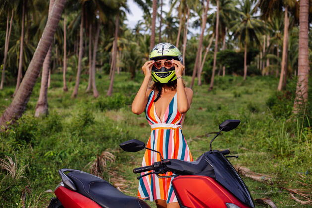 手掌在棕榈树下热带田野的丛林中 穿着黄绿色摩托车头盔和彩色浅夏装的美丽骑手妇女的肖像漂亮光明人