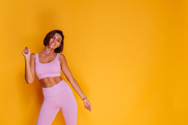 胸部体育女站在黄色的墙上 展示她的腹肌 满意的健身训练和饮食的结果 有一个愉快的面部表情 穿着运动上衣和紧身裤腹肌成人运动员