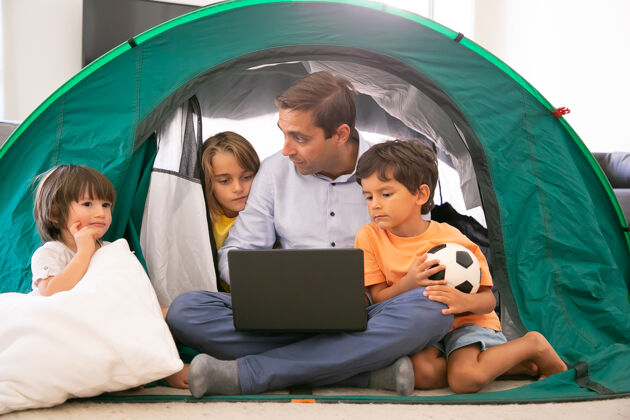 女儿体贴的爸爸盘腿坐在家里的帐篷里 抱着笔记本电脑和孩子们可爱的孩子们和白种人的爸爸在便携式电脑上看电影童年 家庭时间和周末的概念爸爸盘腿爸爸