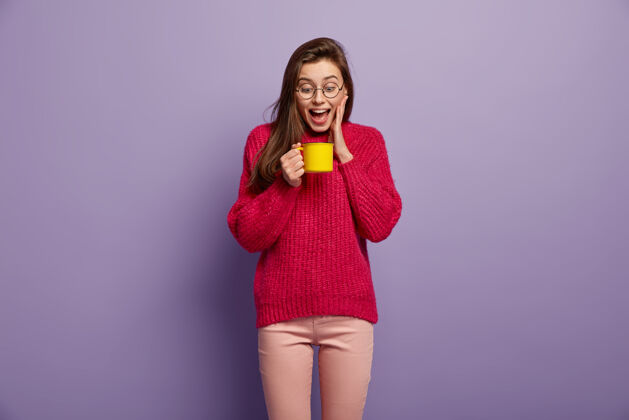 毛衣快乐女生看一杯热腾腾的芳香饮料 拿着黄色的杯子 戴着眼镜 红色的套头衫 靠着紫色的墙站着快乐女生有咖啡休息喝酒的概念浓缩咖啡白种人杯子