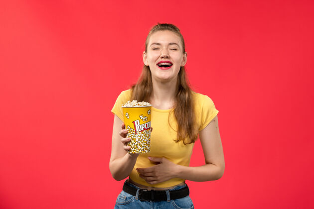年轻的女性正面图年轻女性在电影院拿着爆米花包 笑着看红墙电影院的女性趣味电影漂亮年轻电影院